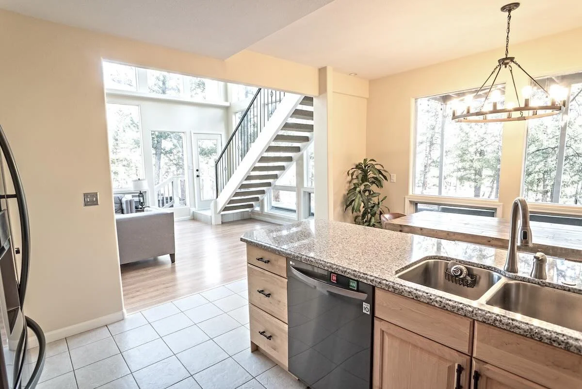Open-concept kitchen and living room area with large windows, kitchen island with sink, dishwasher, granite countertops, and wooden cabinets, leading to a bright living space with stairs and many windows.