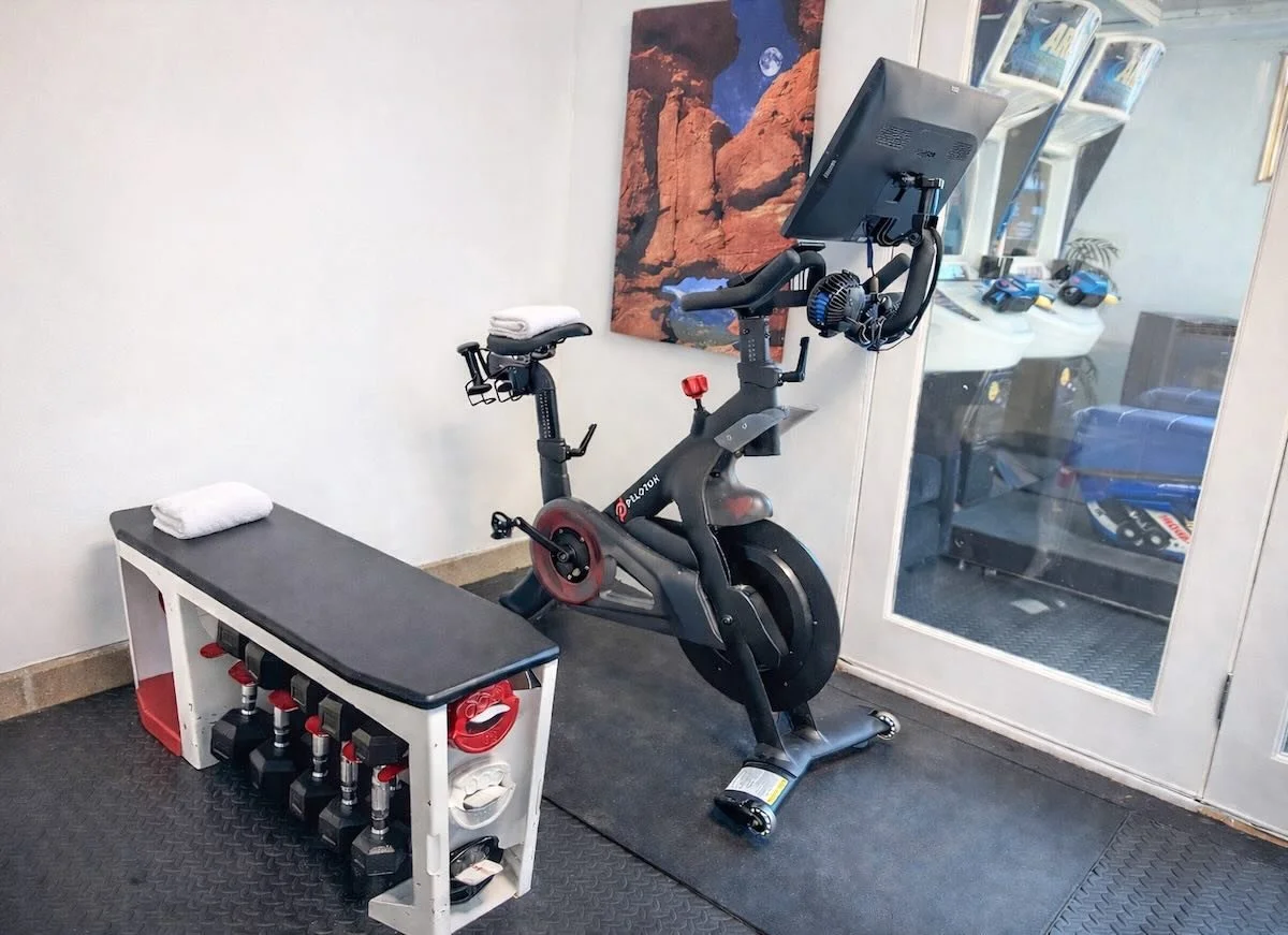 Indoor gym corner with a stationary exercise bike, a white bench with fuel bottles beneath, and a picture of red rock formations and a mountain on the wall.