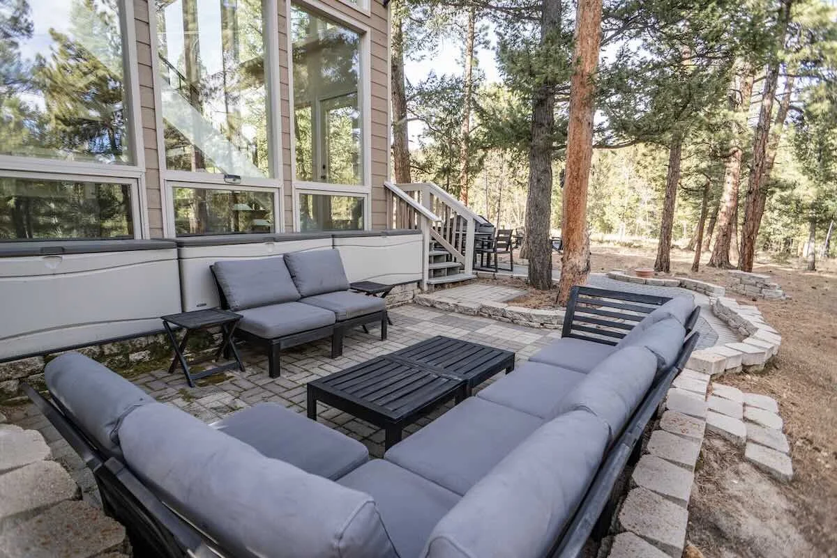 Backyard patio with outdoor furniture, including a sectional sofa and chairs with cushions, surrounded by trees and a forest backdrop.