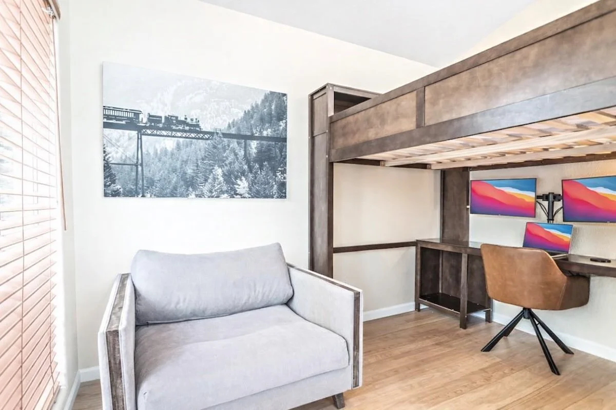 Room with a loft bed, a desk with three monitors, a brown swivel chair, a gray sofa, a framed black-and-white train and mountain photo, and wooden blinds on the window.