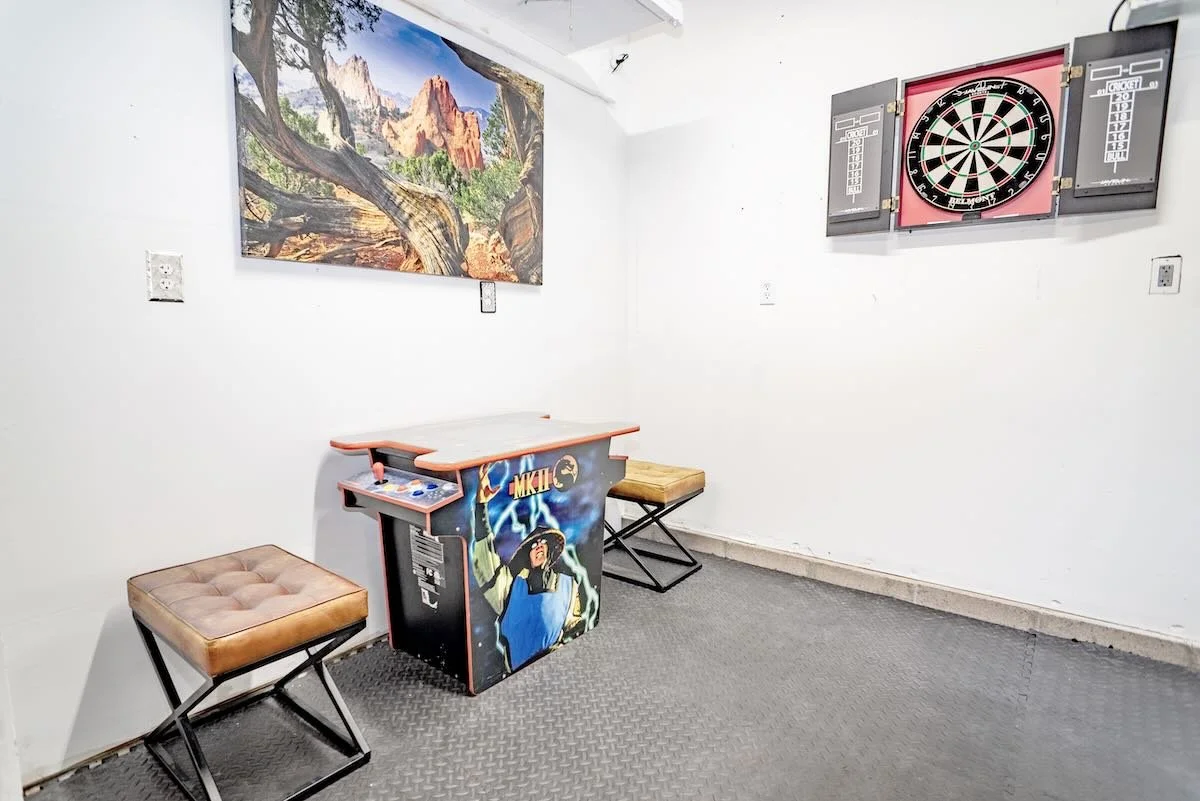 Room with a dartboard on the wall, a small arcade game with a colorful design, two cushioned stools, a large landscape painting, and a gray rubber floor.