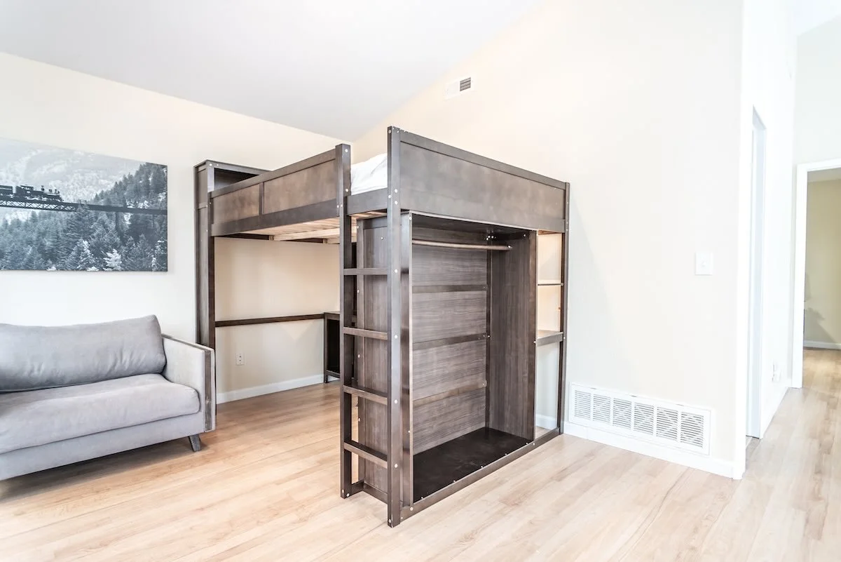 Empty loft bed with gray wood frame in a bright living room, next to a gray sofa and window with natural light.