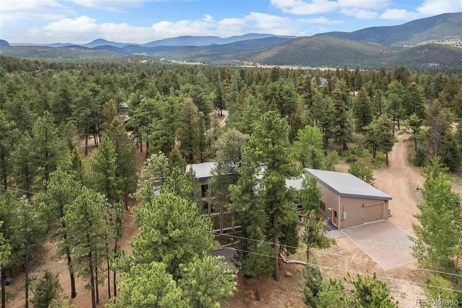 Aerial view of a forested area with pine trees, a small house, and a large building with a garage, with mountains in the background.