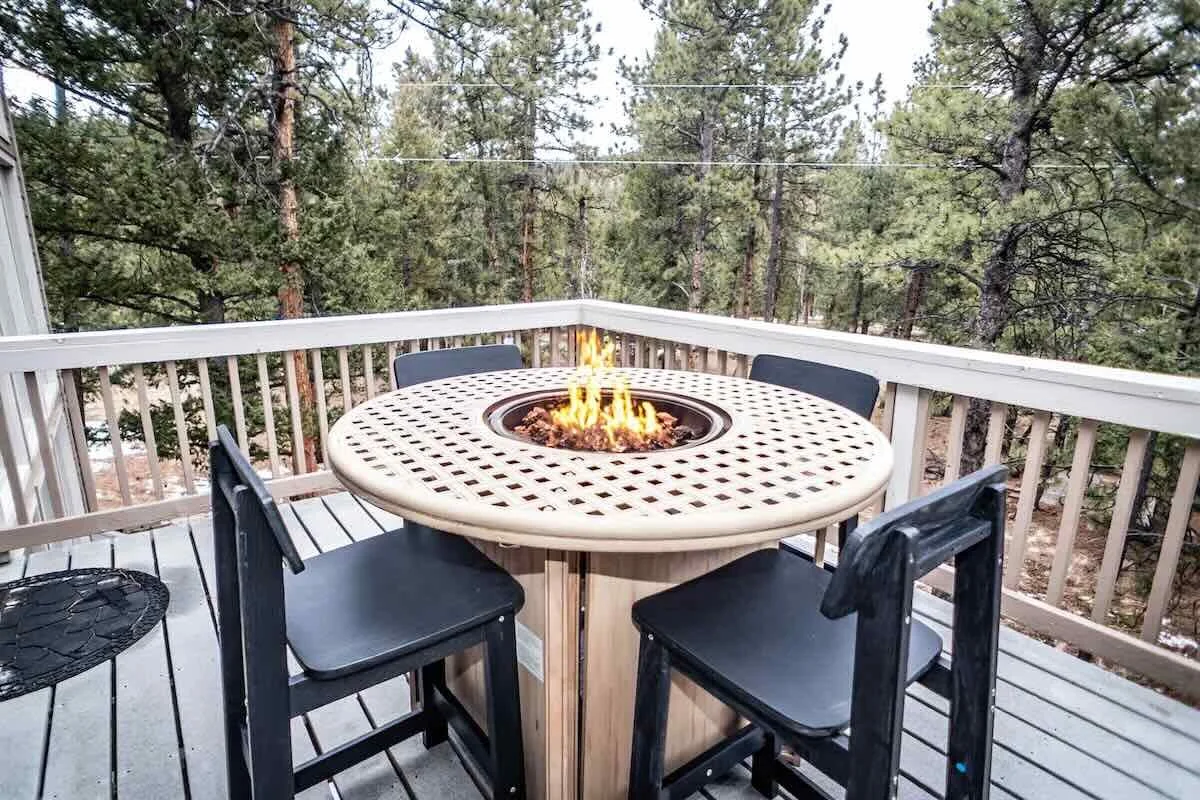 A round outdoor table with a built-in fire feature, surrounded by four black chairs, on a wooden deck with a railing, overlooking a forested area.