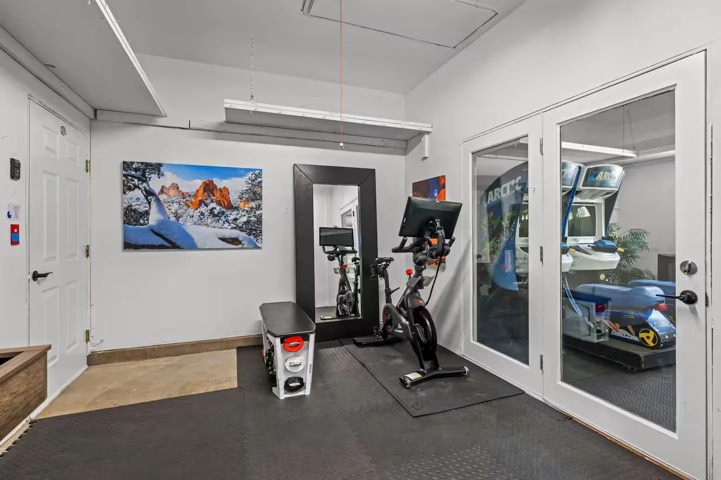 Small indoor gym area with exercise bike, small workout bench, and TV monitor. Behind a glass wall, there are arcade racing games and a foosball table.