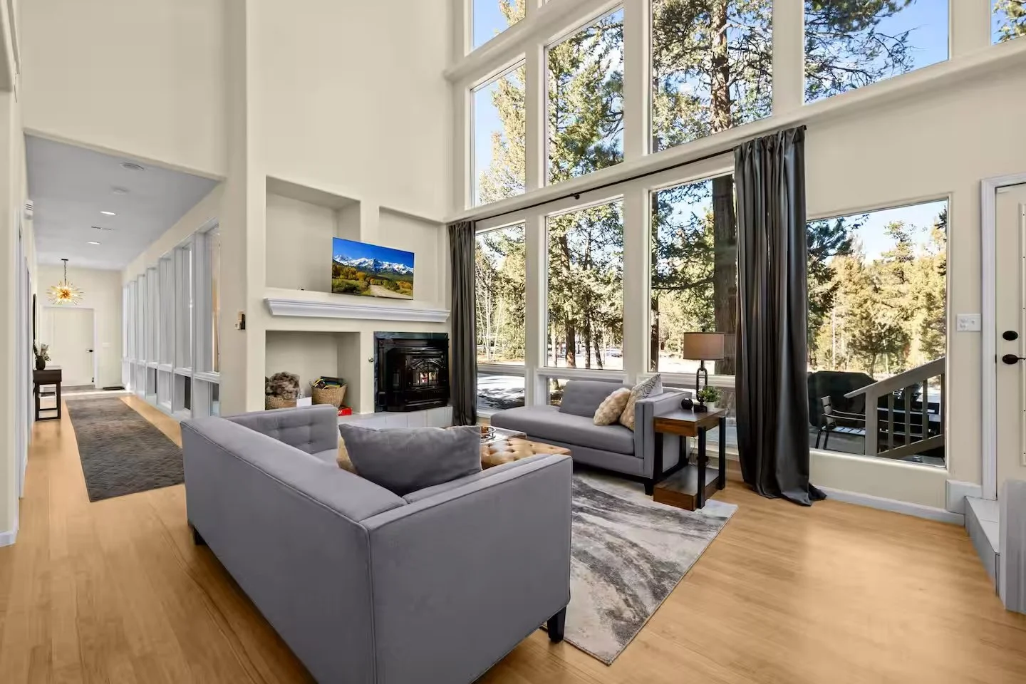 Bright living room with large windows showing trees outside; two light gray sofas with pillows, a coffee table, and a dark wood side table with a lamp; a wall-mounted TV above a fireplace; hardwood floors and curtains framing the windows.