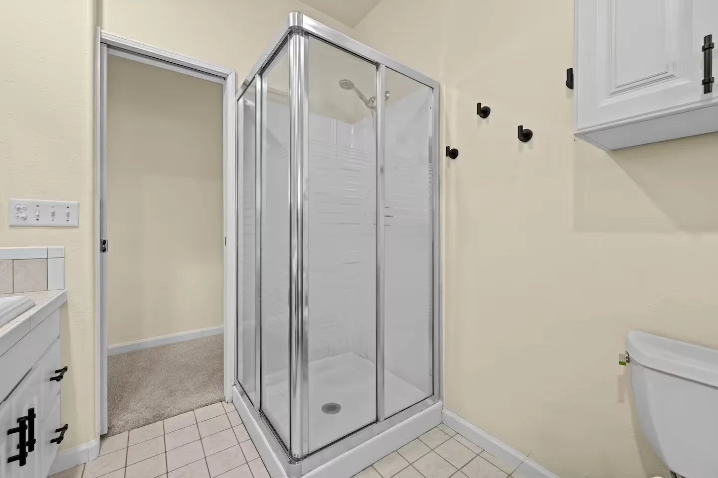 A bathroom corner with a glass shower enclosure, white vanity, and a toilet, with black hooks on a light yellow wall.