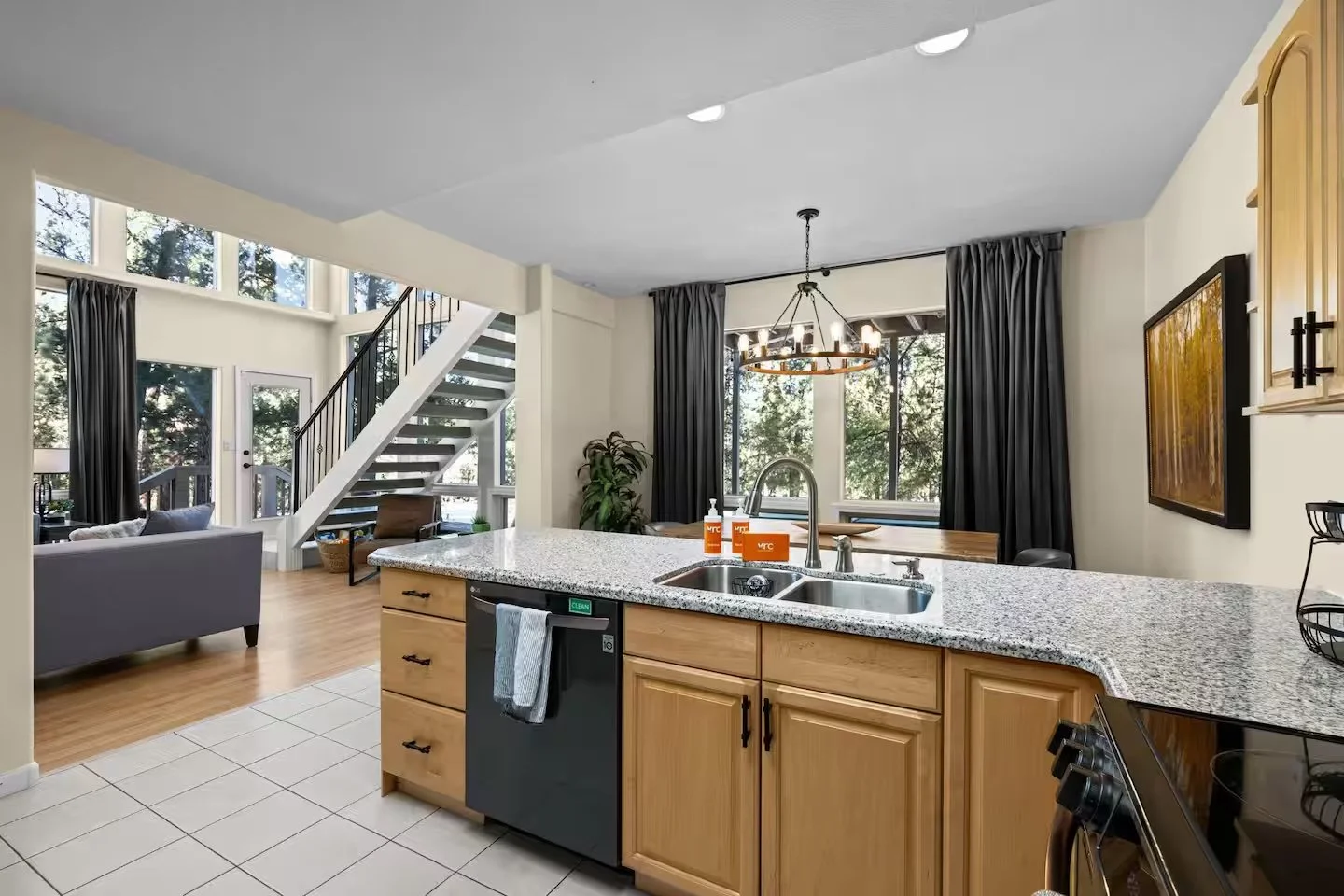 Open-concept kitchen and living area with granite countertops, wooden cabinets, a kitchen sink, and appliances, with large windows, black curtains, a dining table, chandelier, and a staircase leading upstairs.