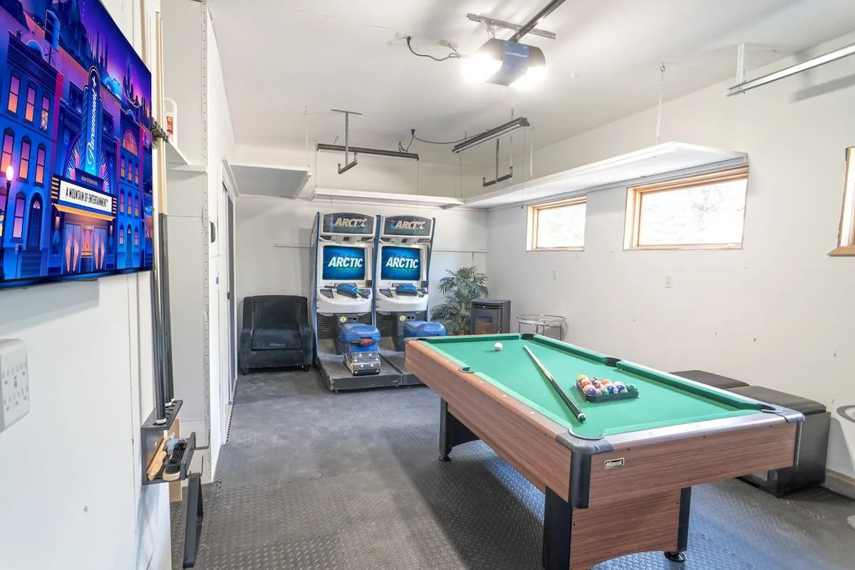 A game room featuring a pool table with billiard balls and a cue, arcade machines labeled 'ARCTIC', a flat-screen TV on the wall, and two small windows letting in natural light.