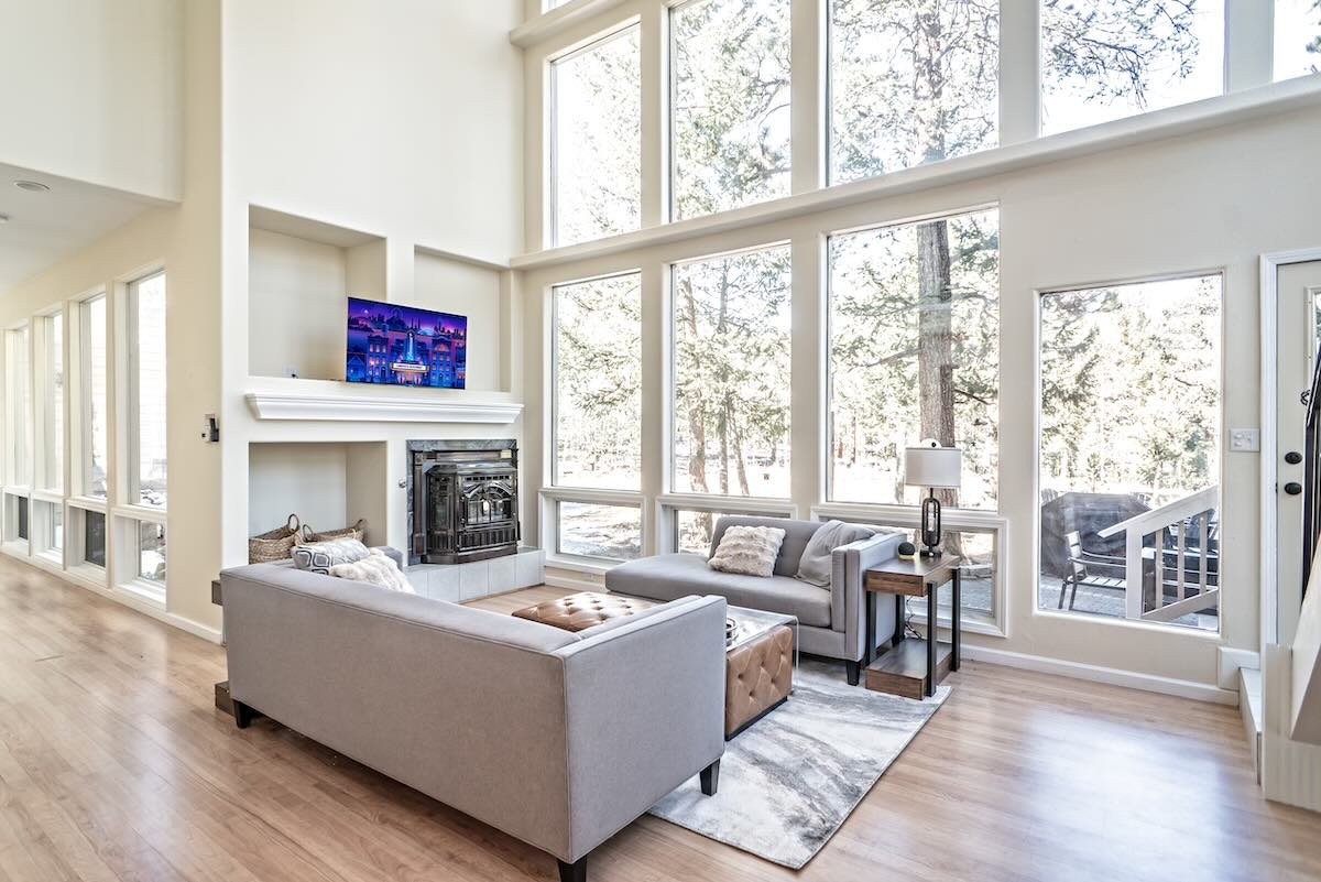 Modern living room with large windows, two light gray sofas, a white fireplace, a mounted TV, a side table with a lamp, and a patterned rug.