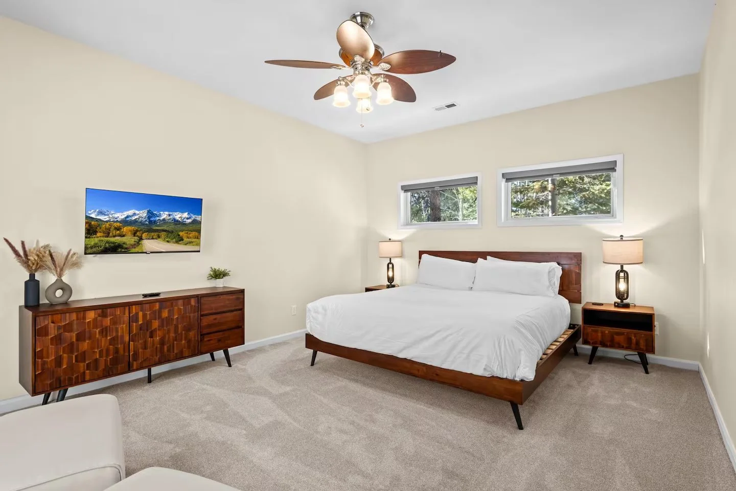 A bedroom with a wooden bed frame and white bedding, two nightstands with lamps, a wall-mounted television, a wooden sideboard, and two narrow windows showing trees outside, with a ceiling fan overhead.