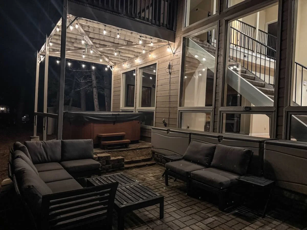Nighttime outdoor patio with string lights, outdoor furniture, hot tub, large windows, brick flooring, and an exterior staircase.