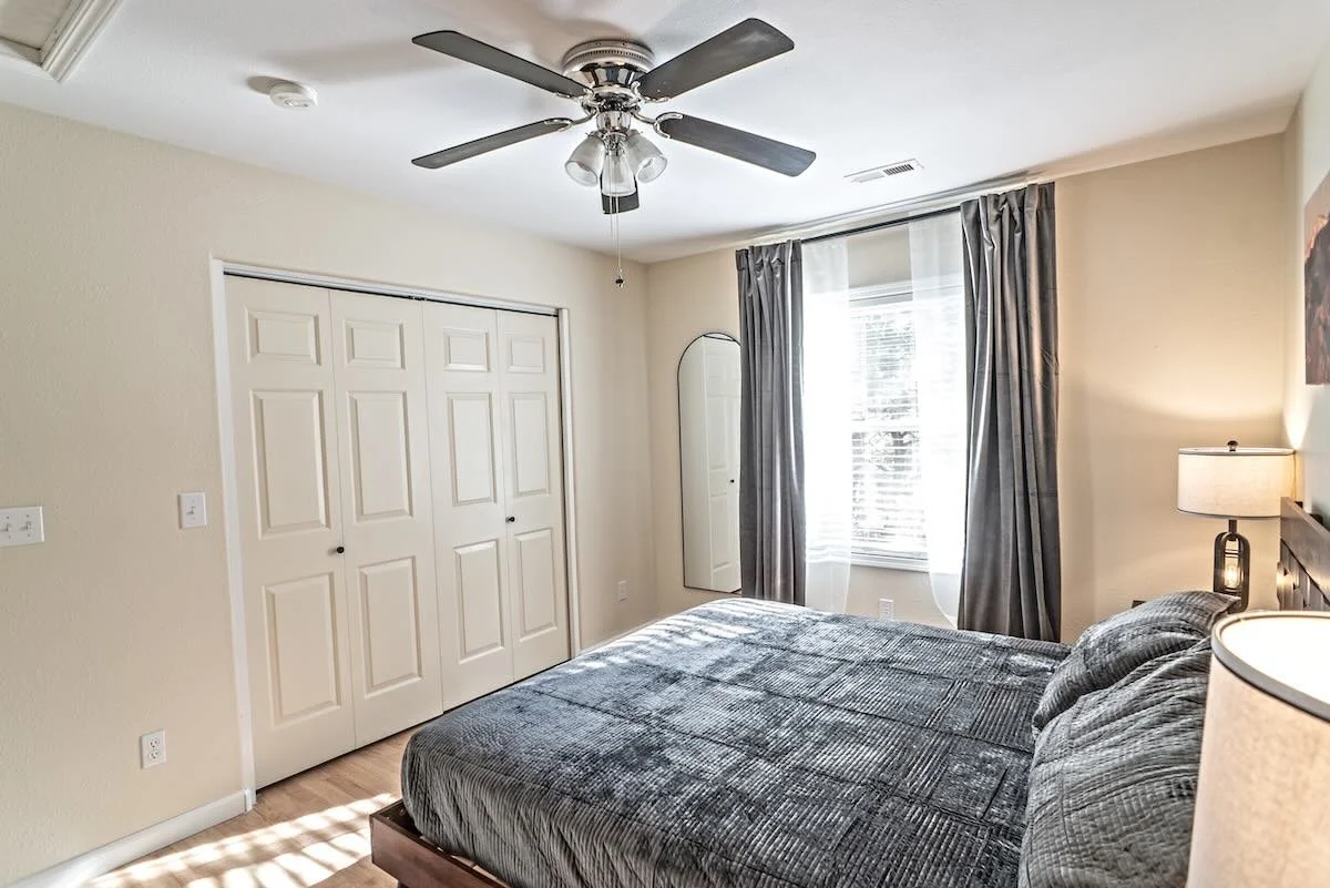 A bedroom with a bed, a window with curtains, a ceiling fan, a lampshade, and a closet with two doors.