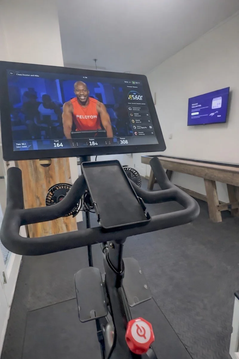 A stationary exercise bike in a room with a large screen displaying an indoor cycling workout, showing a smiling instructor in a red shirt and workout stats.
