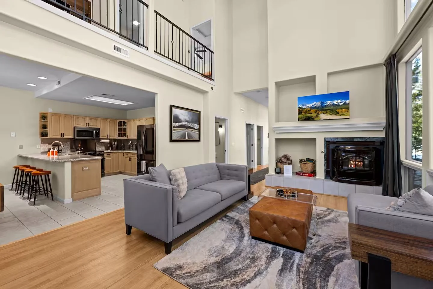 Living room with high ceiling, light-colored walls, large windows with curtains, a gray sofa, a wooden coffee table, fireplace, and three potted plants in the background. Adjacent to the living room is an open kitchen with wooden cabinets, black appl