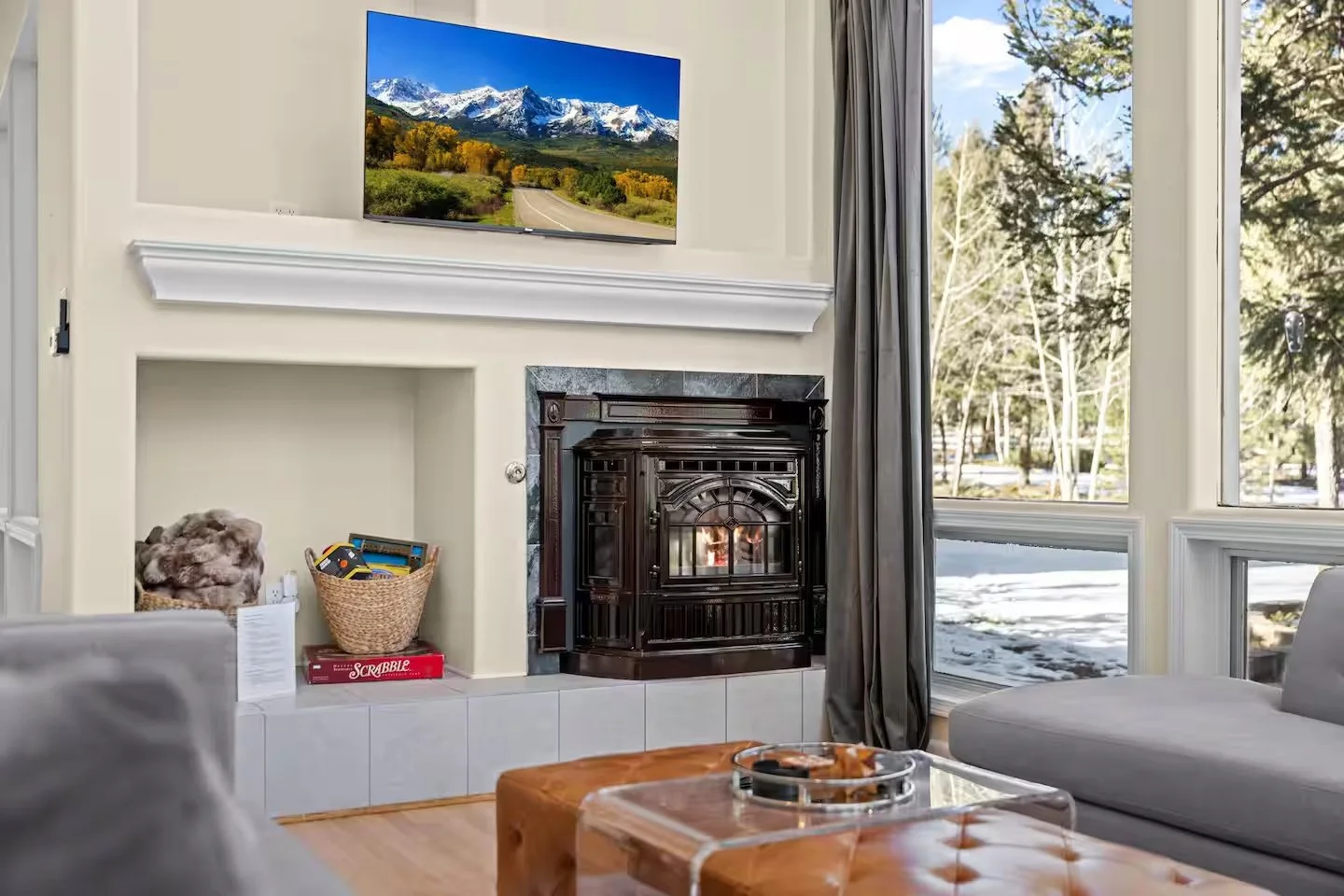 Living room with a fireplace, a flat-screen TV mounted above it, and large windows showing a snowy outdoor scene with trees.