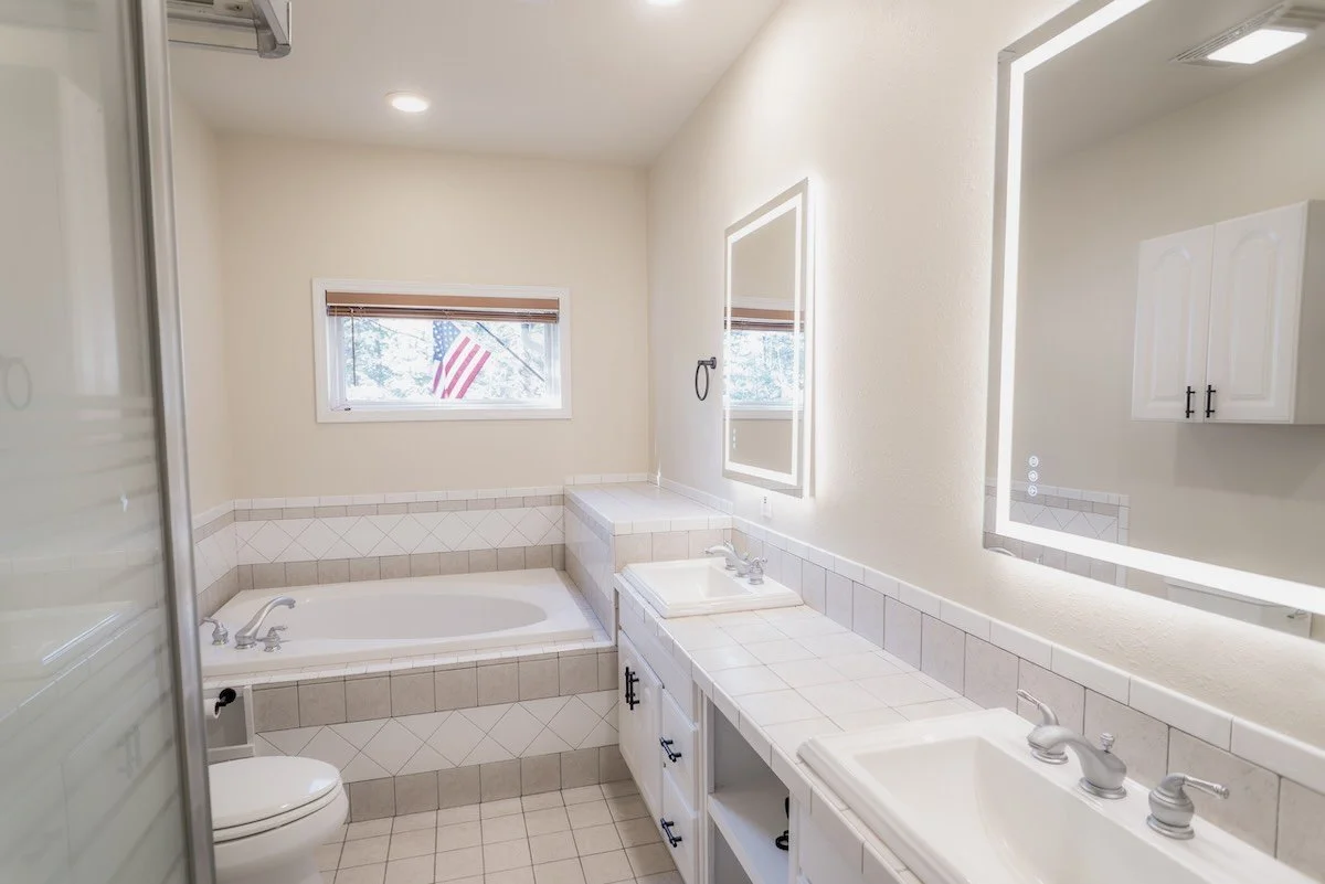 Bathroom with a large bathtub, double sink vanity with mirrors, window with an American flag outside, white tile surfaces, and a separate shower enclosure.