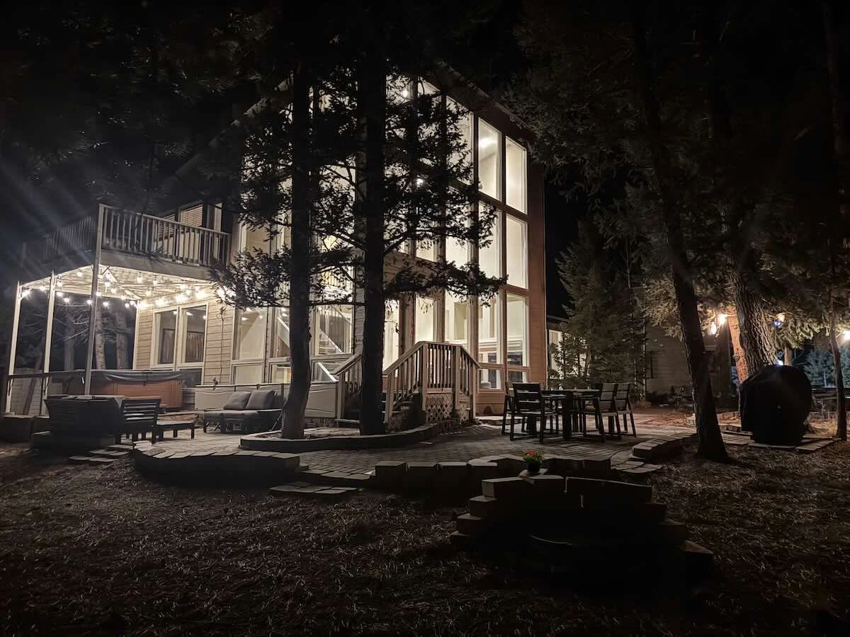 Night view of a house surrounded by trees, with illuminated windows, a backyard deck with outdoor furniture, string lights, and a barbecue grill.