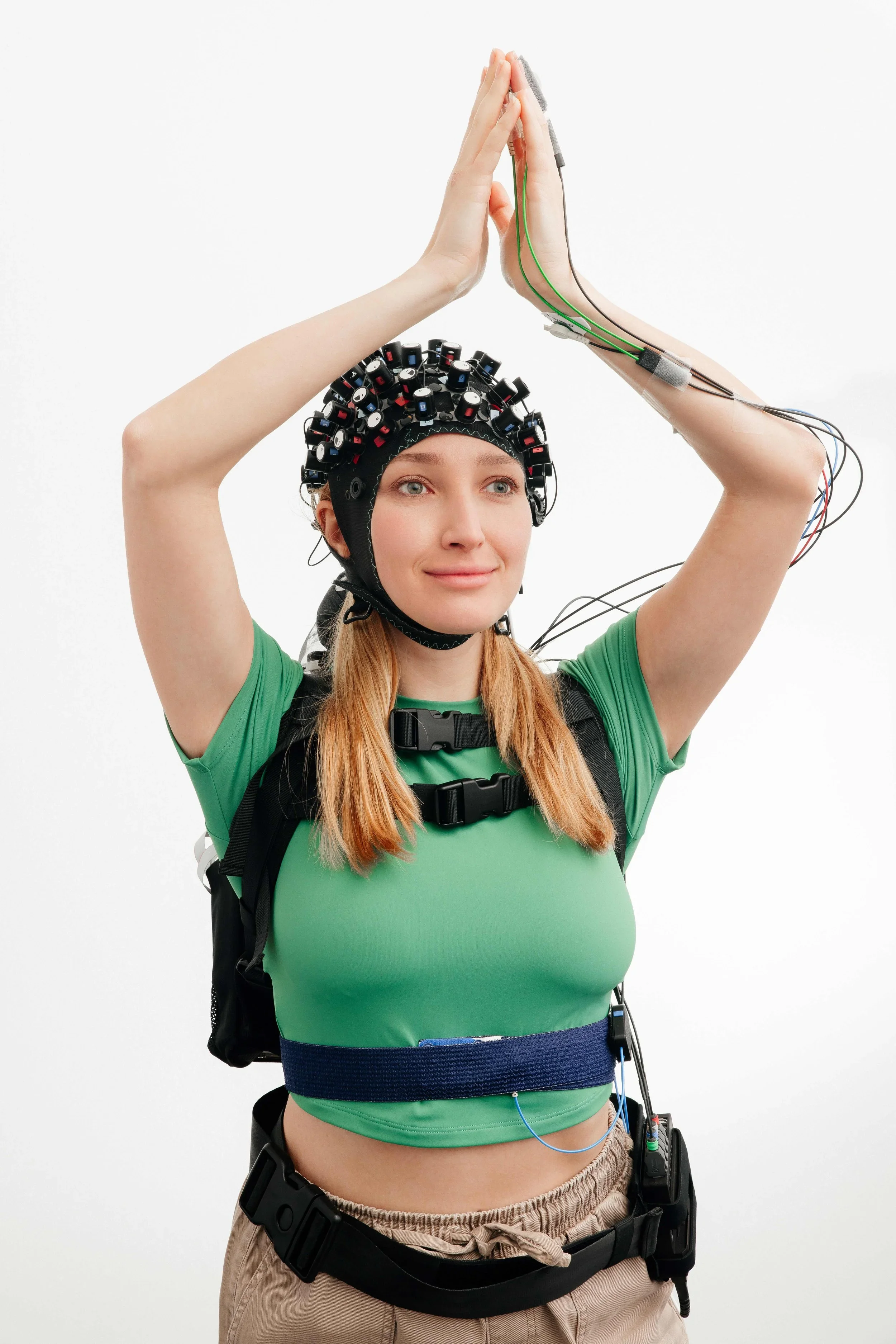 Woman wearing a fNIRS cap with electrodes, connected to wires, standing against a white background. She has a slight smile, with her palms pressed together above her head.