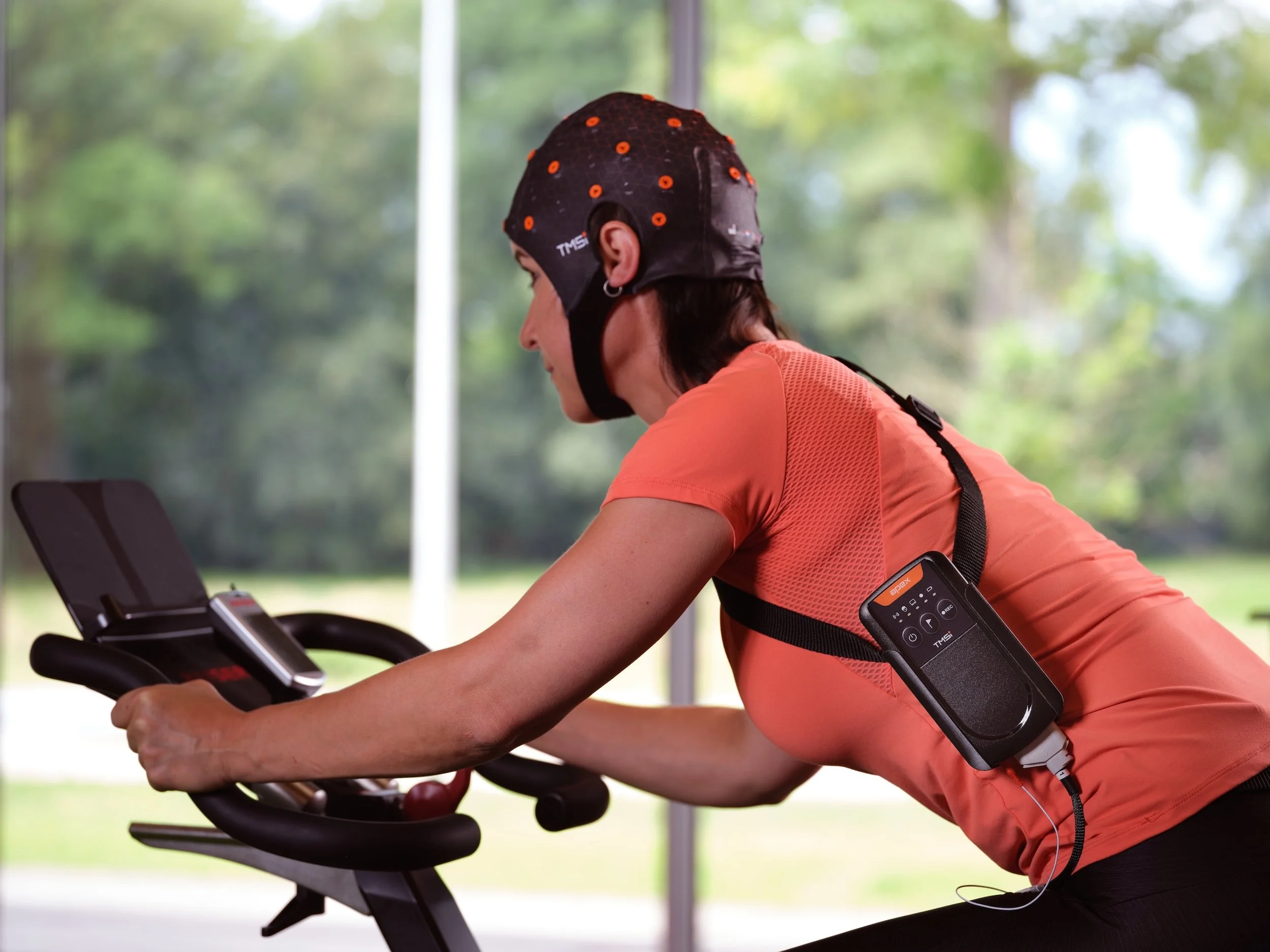Woman exercising on stationary bike, wearing a brain monitoring cap and portable EEG device attached to her chest.