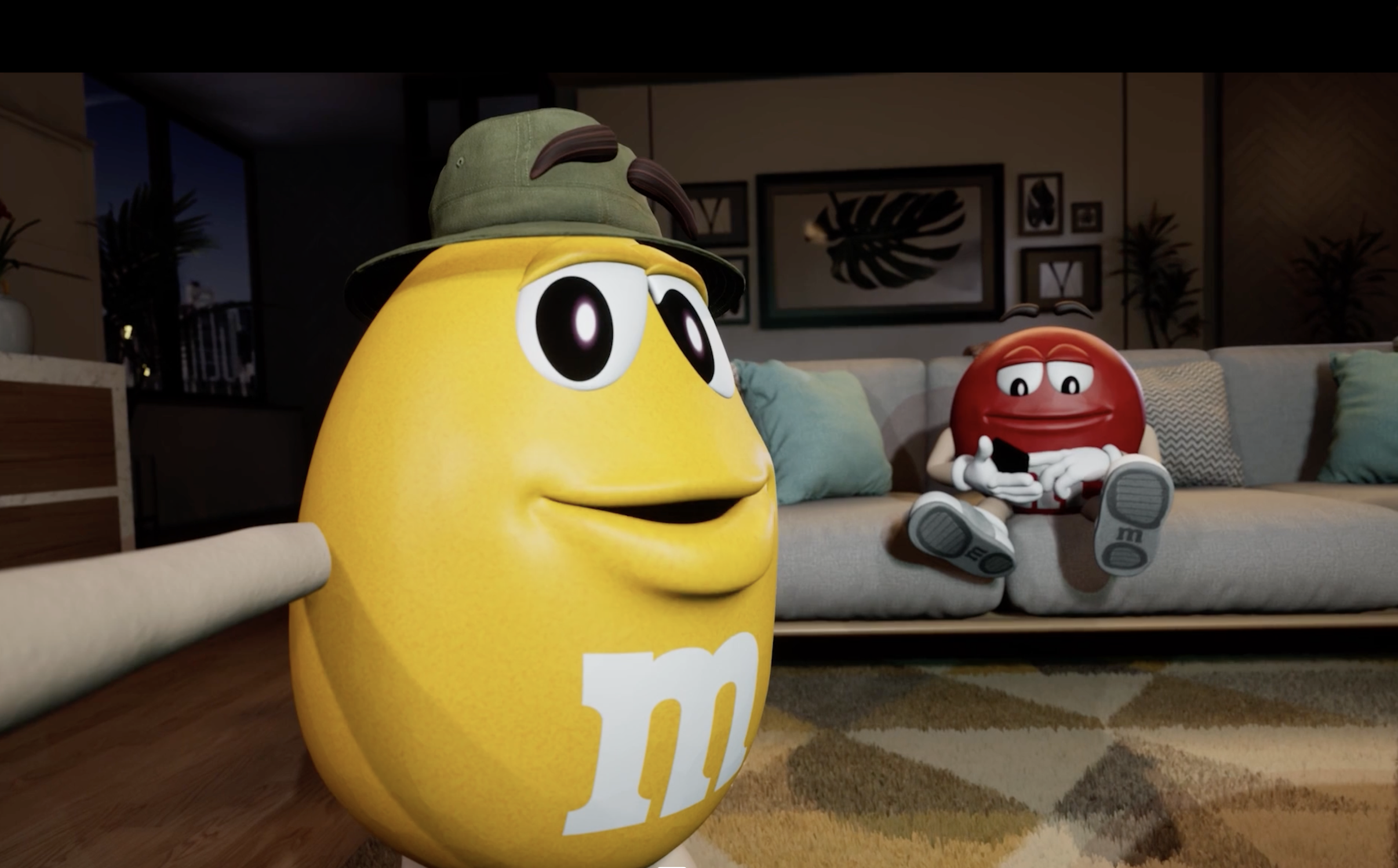 Yellow M&M character wearing a safari hat, standing indoors with a living room background.