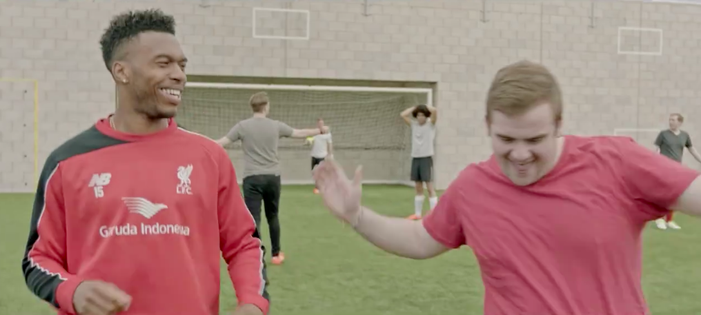 Two men smiling and high-fiving on a soccer field, with other people in the background practicing and preparing for a game.