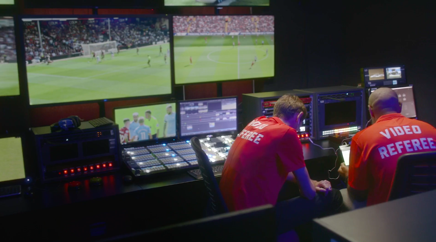Two video referees in red shirts working in a control room with multiple monitors displaying a soccer match.