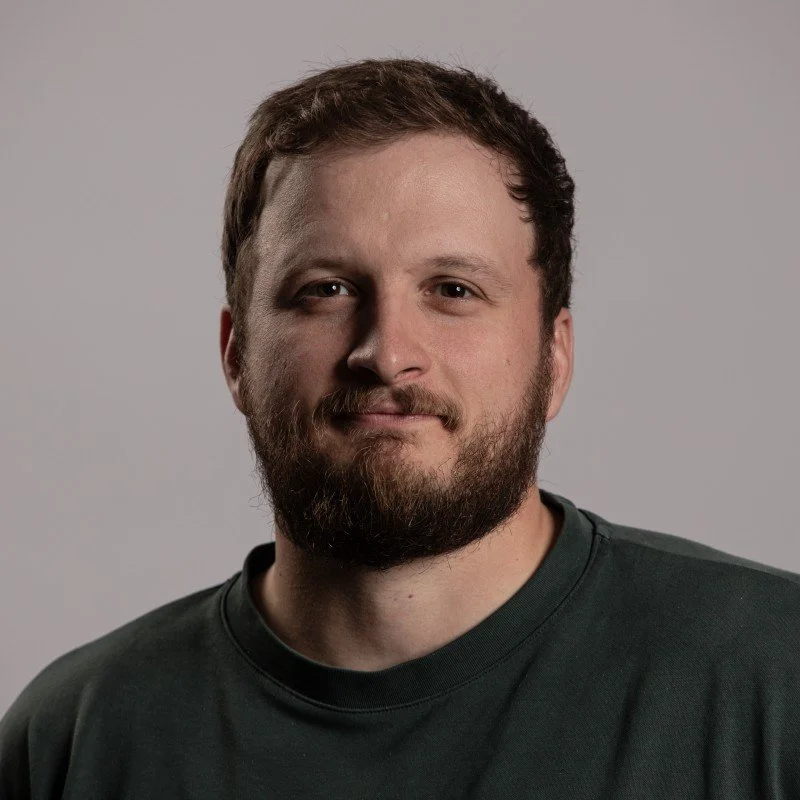A man with a beard and short brown hair, wearing a dark t-shirt, standing against a plain light gray background.