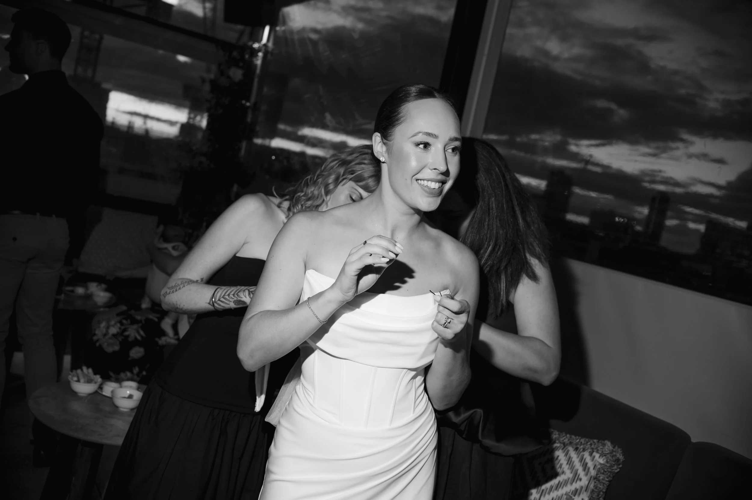 A woman in a strapless white dress smiling and posing, with others behind her, at an indoor gathering during sunset.