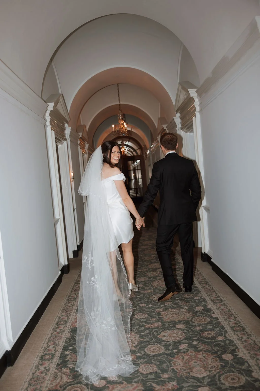 A bride and groom holding hands and walking down a hallway in a wedding venue, with the bride smiling and wearing a white dress with a veil and the groom in a black suit.