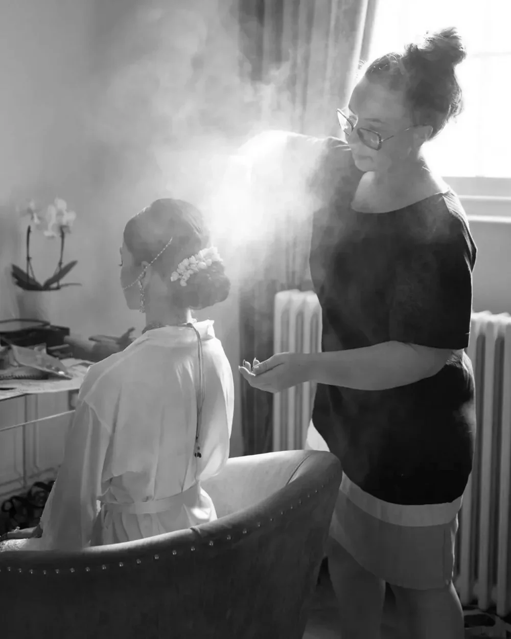 A woman in casual clothing applies makeup or touches up the face of a seated bride, who is dressed in a white robe and adorned with floral jewelry, in a softly lit room.