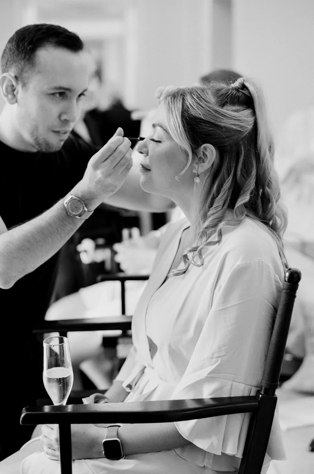 A woman with styled hair and earrings gets her makeup done by a man in a black t-shirt at a makeup station. The woman is seated with a glass of champagne on her lap, wearing a light-colored robe. The scene appears to be in a dressing or makeup room.