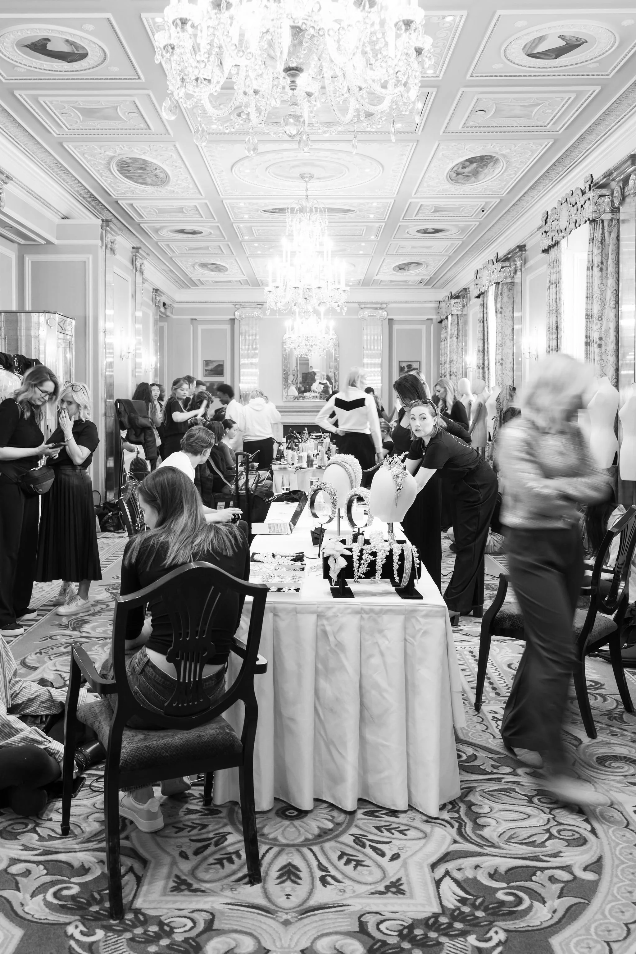 A ballroom decorated with chandeliers and ornate ceiling panels, filled with people attending a jewelry display event. Tables hold jewelry displays, with women browsing and engaging with vendors.