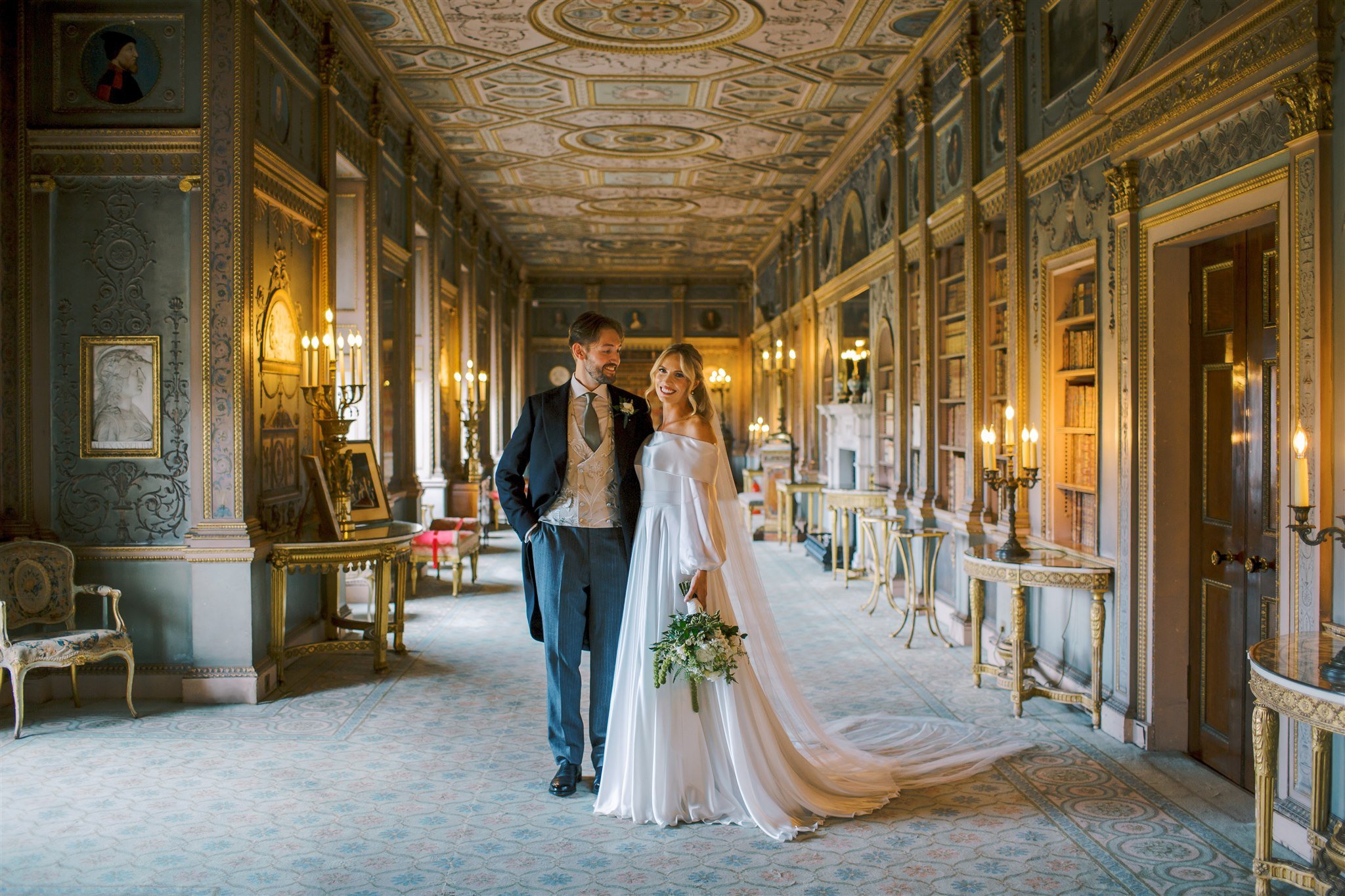 A bride and groom standing together in a grand, ornate hallway with gold accents, intricate ceiling details, and classical decor, sharing a joyful moment on their wedding day.