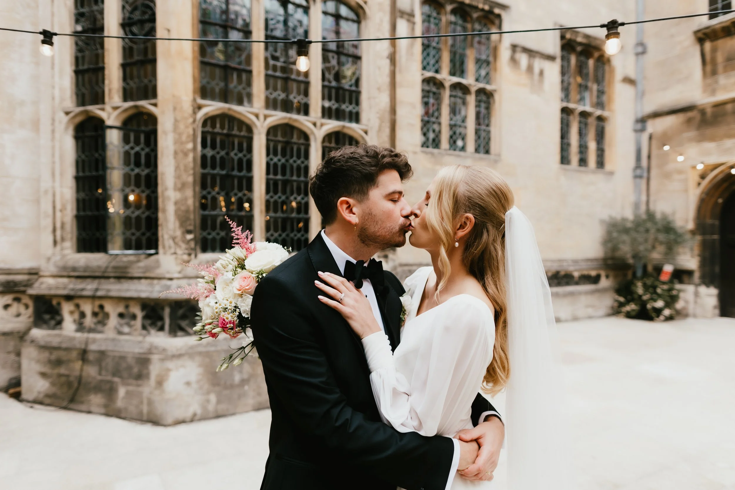A bride and groom kissing in front of a historic stone building with large leaded glass windows, string lights overhead, and a bouquet of flowers.