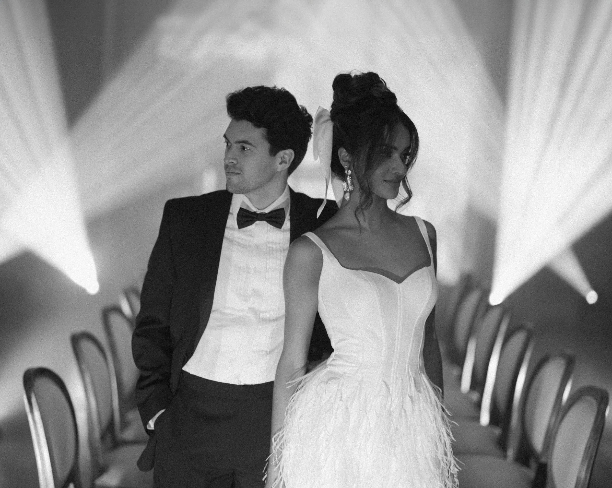 Black and white photo of a man and woman dressed in formal attire, standing with the woman in front of the man, in an elegant setting with chairs and soft lighting.