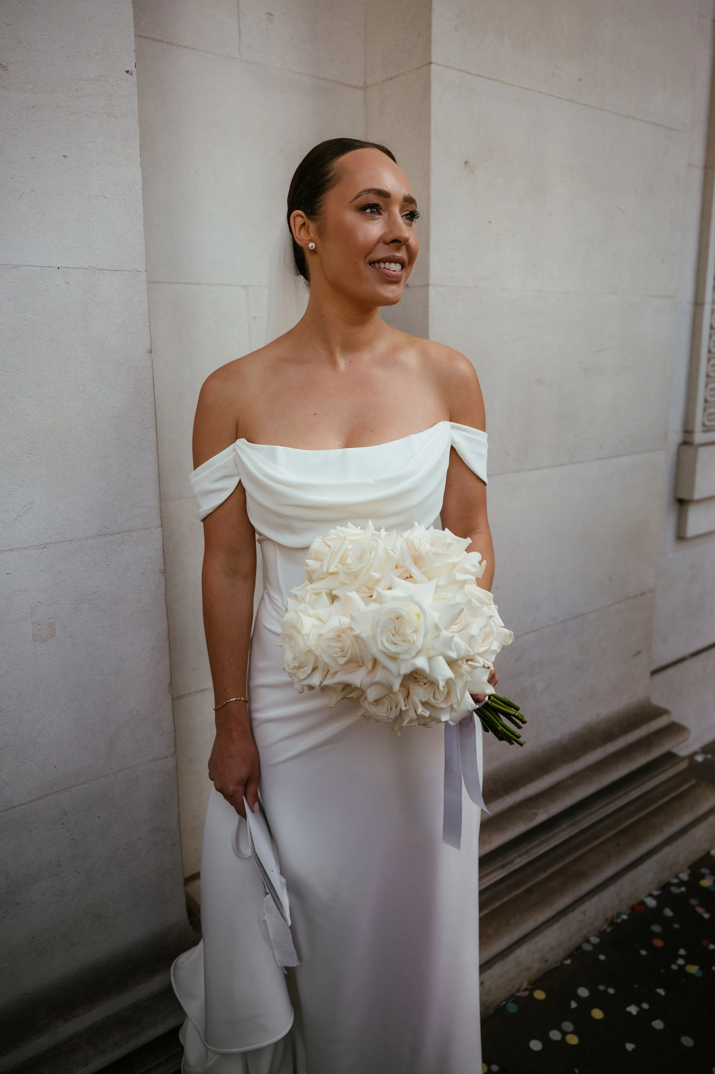 A bride in a white off-shoulder wedding gown holding a bouquet of white roses, standing against a stone wall, smiling.