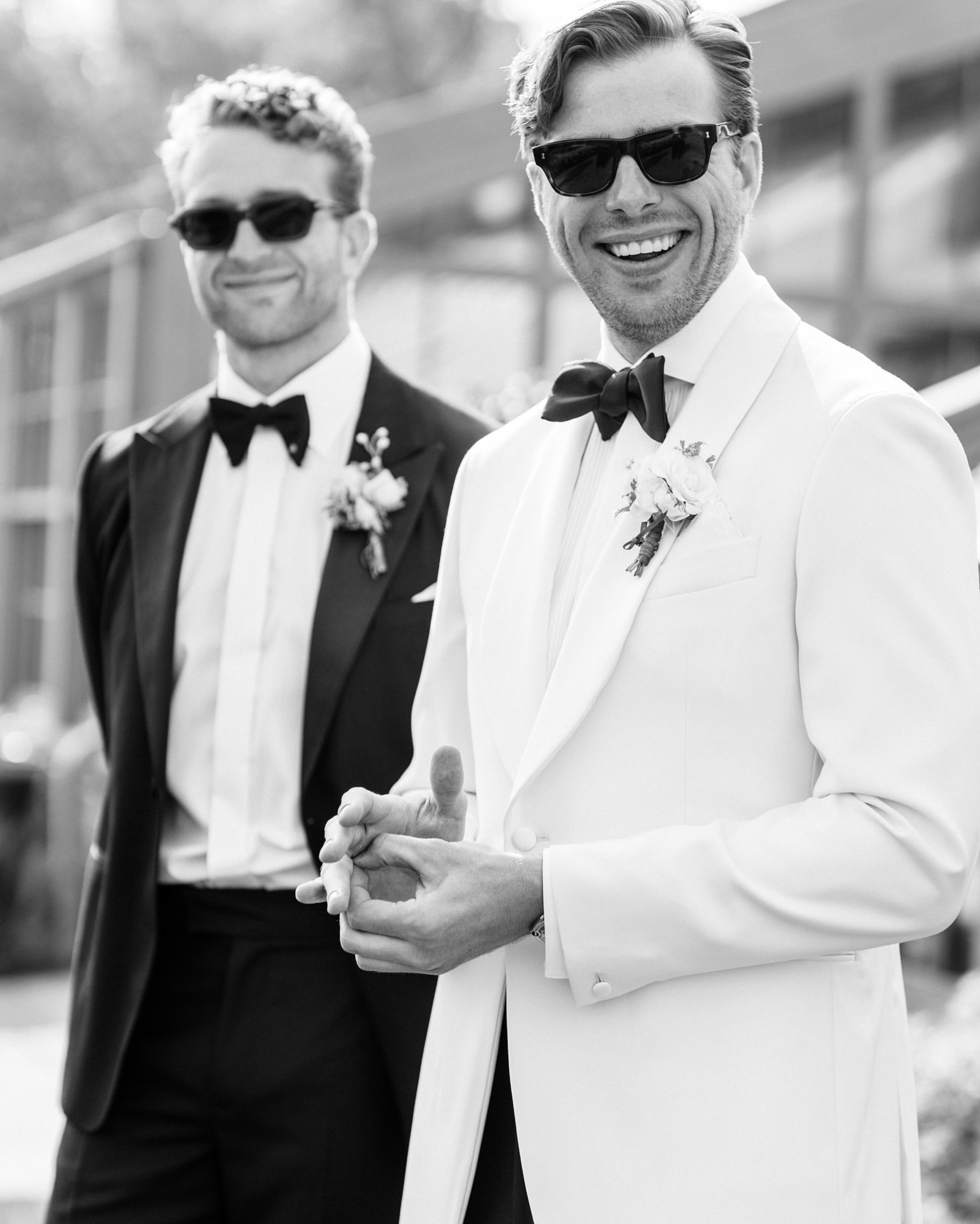 Two men wearing tuxedos, sunglasses, and boutonnières, smiling outdoors at a wedding.