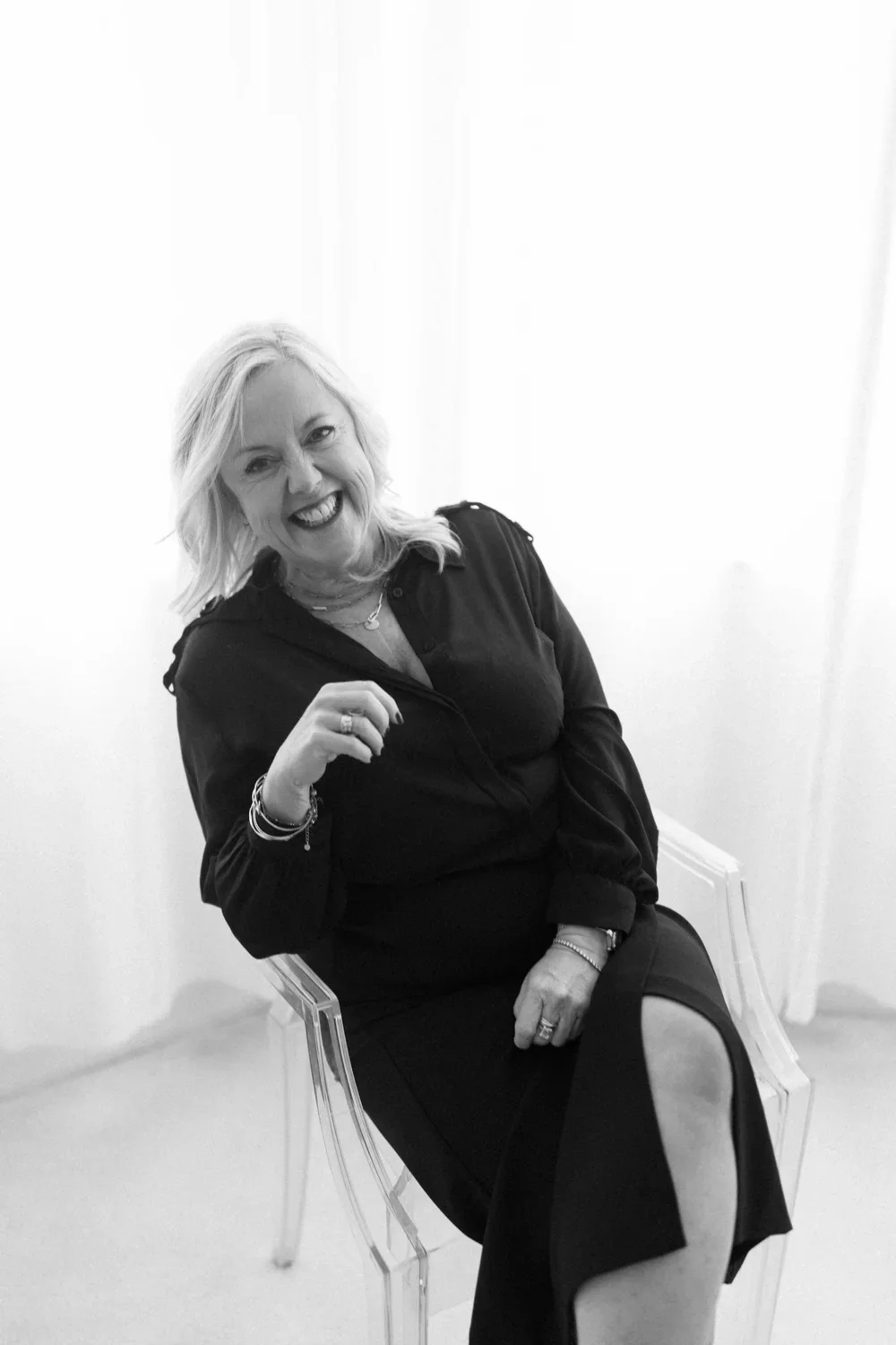 A smiling woman with light-colored hair sitting on a transparent chair in a bright room. She is wearing a dark outfit and jewelry, and gesturing with her right hand.