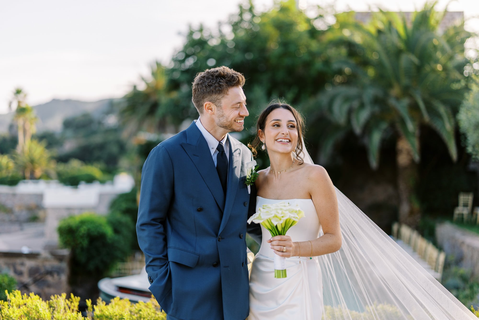 A newlywed couple standing outdoors, smiling at each other, with the bride holding a bouquet of white calla lilies. The groom is in a navy blue suit and the bride is in a strapless white wedding dress with a long veil.