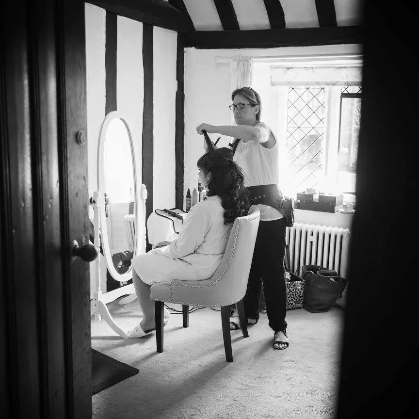 A woman getting her hair styled by a hairstylist in a room with a mirror, window, and various hairstyling tools.