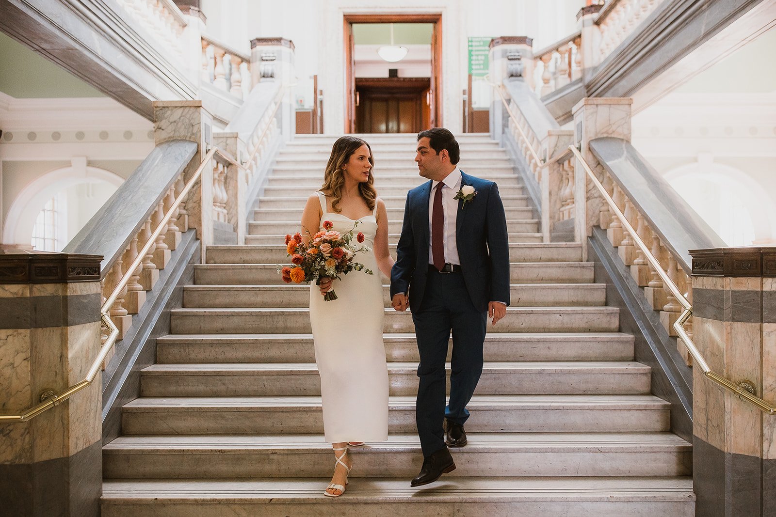 A newlywed couple walking down a grand staircase in a historic building, holding hands. The bride is holding a bouquet of flowers, and the groom is wearing a suit with a boutonnière.