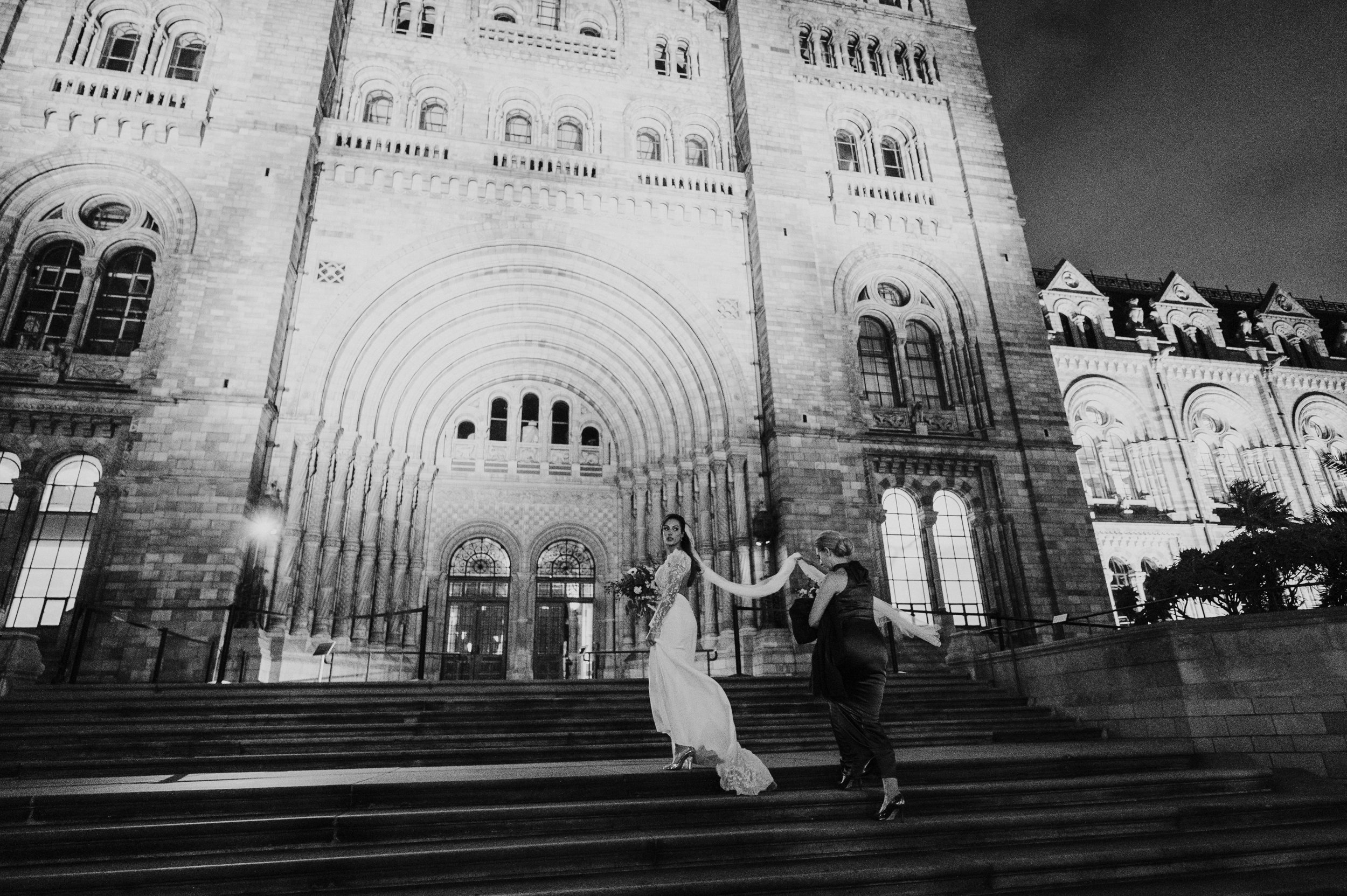 A black and white photo of two women on steps in front of a large historic building at night. One woman is in a white gown holding a bouquet, and the other woman is in a dark dress. They are holding hands and dancing.