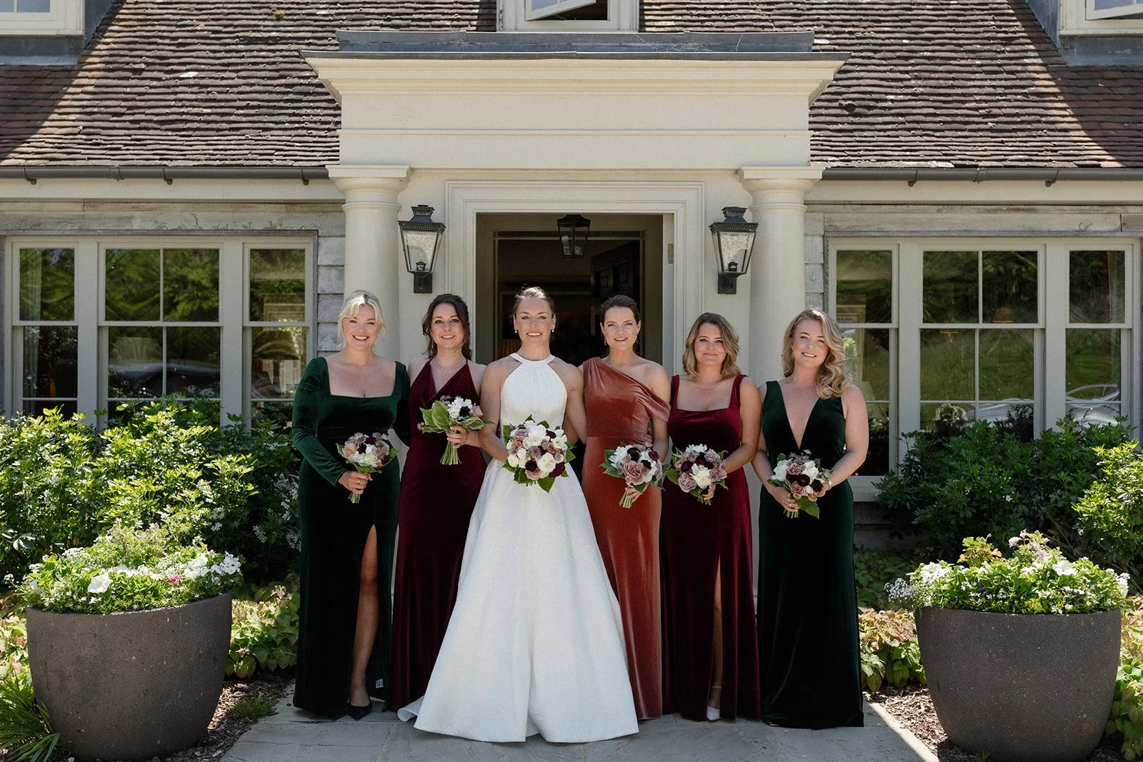 Group of women in elegant dresses holding bouquets, standing in front of a house with large windows and a garden.