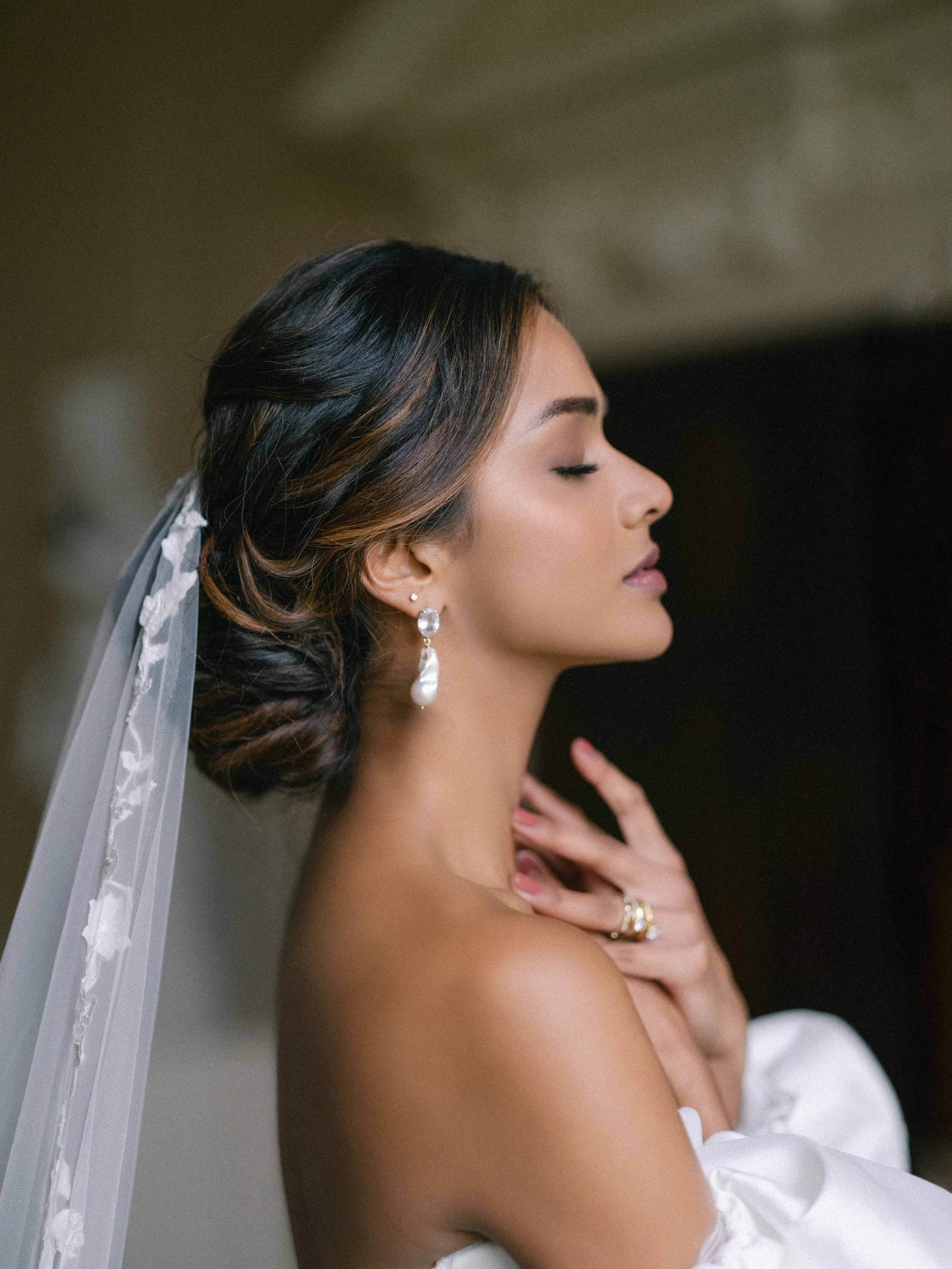 Close-up side profile of a bride with closed eyes, wearing pearl and gemstone earrings, with her hair styled in an elegant updo and a white wedding dress, against a blurred background.