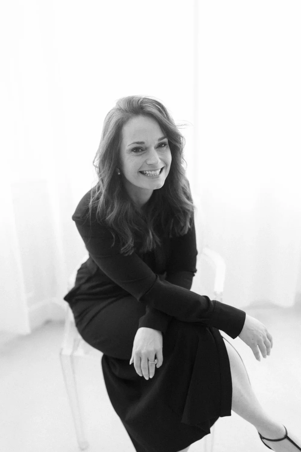 A smiling woman with wavy hair in a black dress sitting on a translucent chair in a bright room.