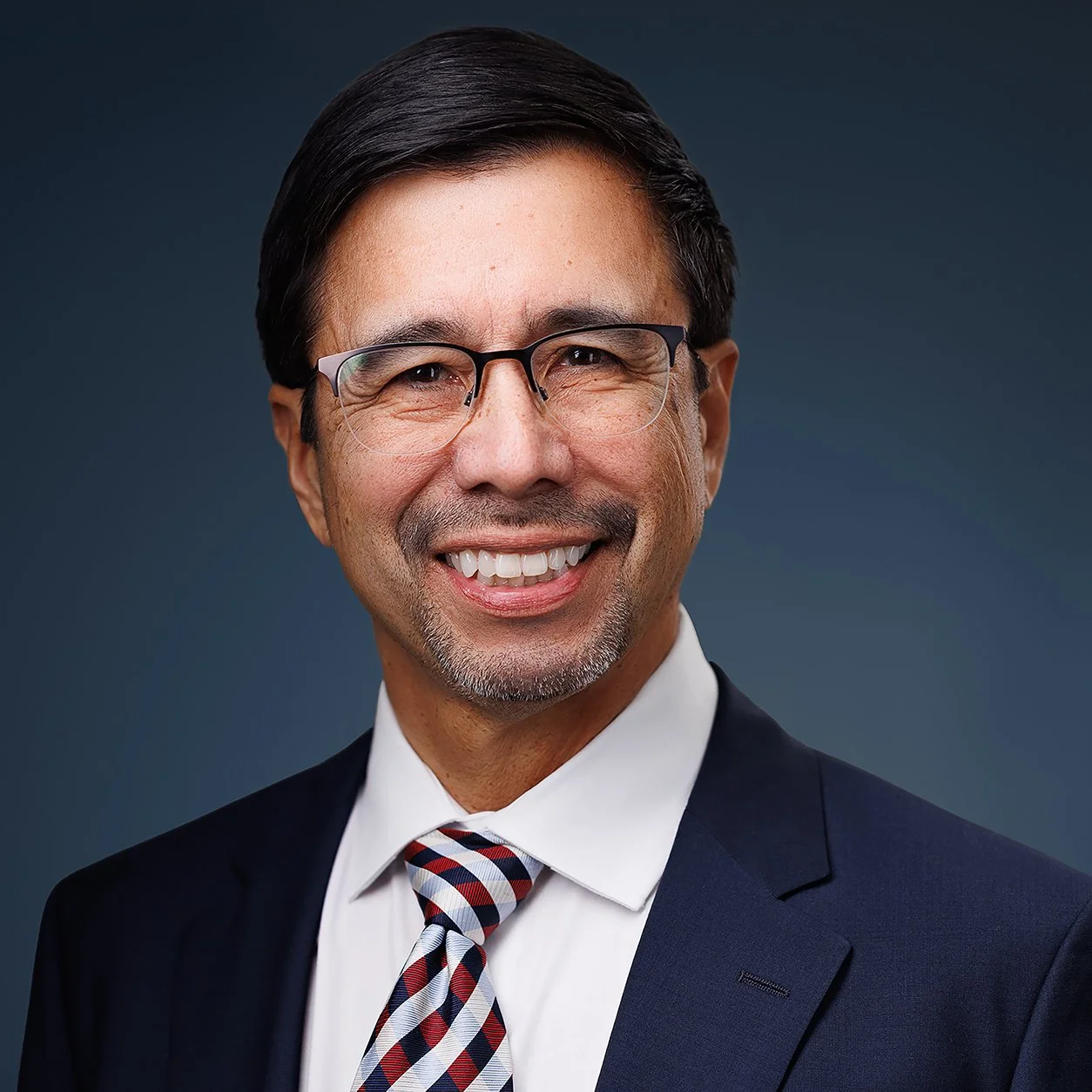 Professional headshot of a smiling middle-aged man with dark hair, glasses, wearing a navy suit, white shirt, and a patterned tie, against a dark gray background.