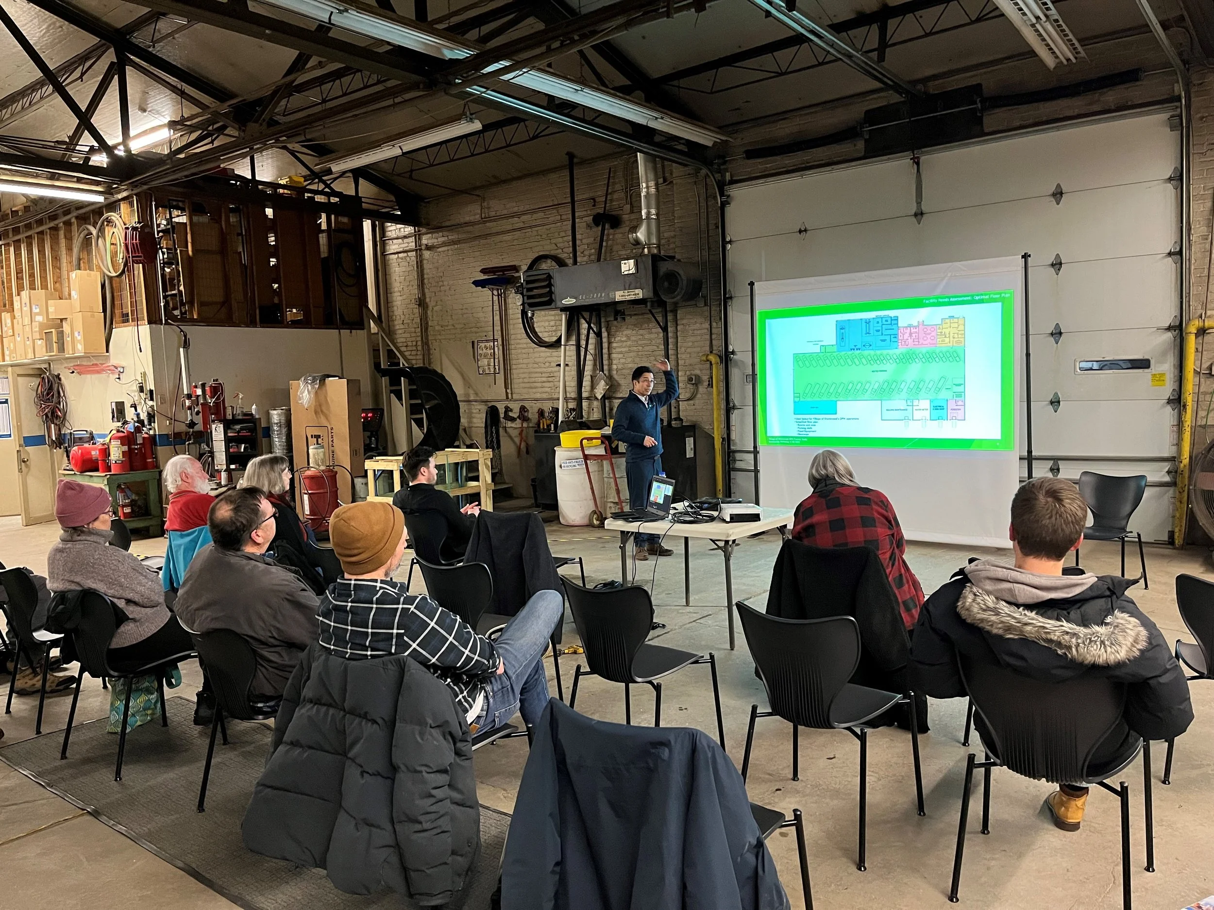A group of people seated in a garage or warehouse, attending a presentation by a man standing near a projection screen displaying a colorful diagram or map.
