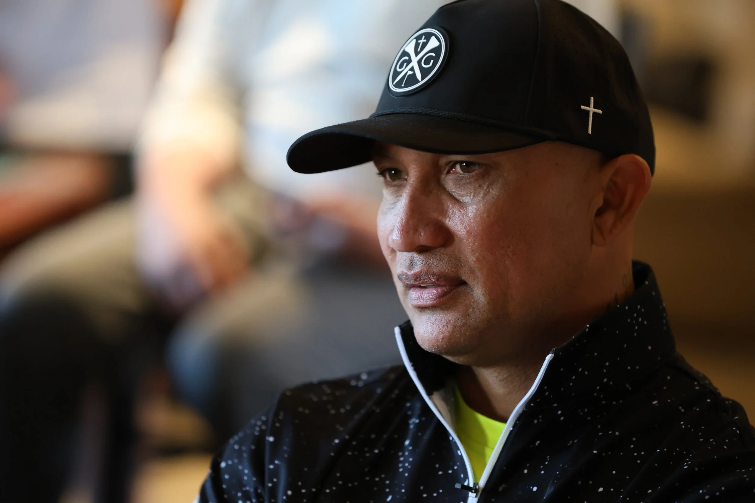 A man wearing a black baseball cap with a religious symbol and a black sports jacket with white dots. He is looking to the side with a serious expression.