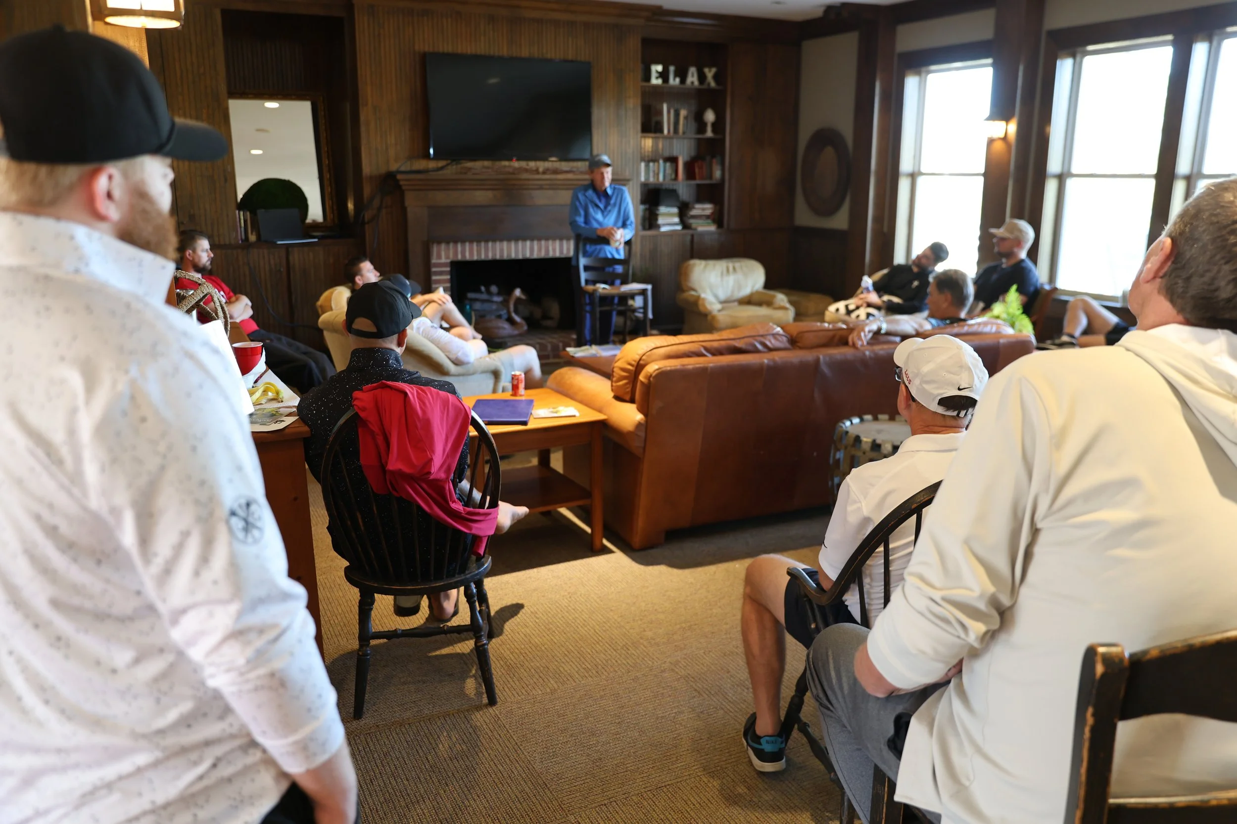 A group of people gathered in a cozy living room with wood-paneled walls and large windows, listening to a man speaking at the front near a fireplace.
